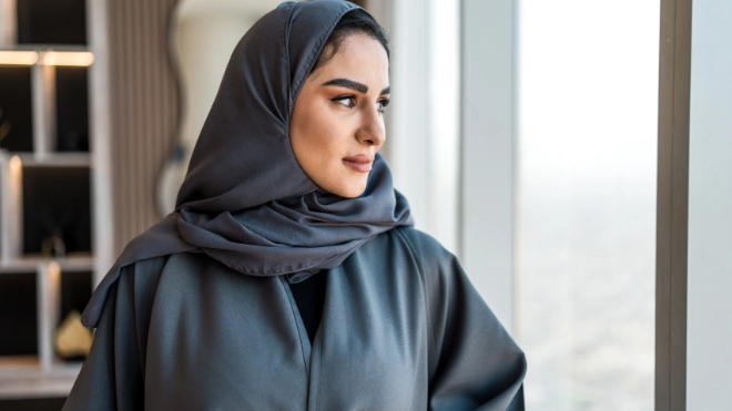 An Arabic woman wearing a gray abaya and matching hijab looks thoughtfully out of a large window with a city view.
